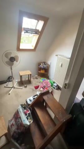 The video pans across a cluttered attic room. A fan is positioned near a small wooden stool, with various items scattered on the floor. A slatted wooden structure, possibly a makeshift ladder or shelving unit, is in the foreground. Beyond this, an open doorway reveals a darker room with wooden flooring and two windows. The walls are painted white, and a radiator is visible. The overall impression is one of disarray and unfinished space.