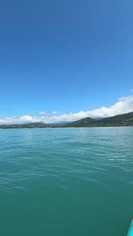 The video shows a first-person perspective of someone on a paddleboard on a bright, sunny day. The water is a clear turquoise color, with gentle waves. In the distance, a coastline of green, tree-covered hills stretches across the horizon, meeting a bright blue sky dotted with fluffy white clouds. The front of a teal paddleboard and a paddle are visible in the lower right corner of the frame. The camera is tilted upwards, emphasizing the vastness of the sky.