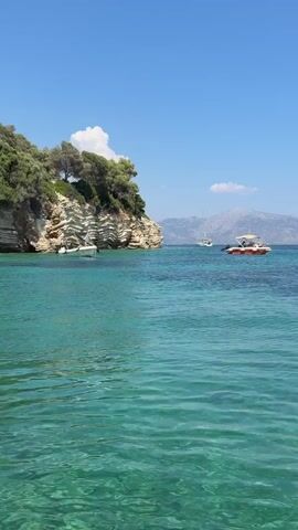 A calm, clear turquoise sea stretches across the foreground of the image. In the middle ground, several boats are visible: a small white boat is anchored near a rocky, tree-covered cliff face on the left. Further out to the right, a sailboat with a white sail is cruising, and a smaller red and white speed boat is also moving across the water. The background features a hazy mountain range under a bright blue sky with a few scattered white clouds. The overall scene depicts a serene coastal environment on a sunny day.