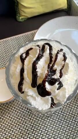 The video shows a close-up of a dessert served in a clear, stemmed glass. The dessert appears to be a milkshake or a similar blended drink, topped with a generous amount of whipped cream and drizzled with chocolate syrup. The glass sits on a gray and black woven placemat on a table. A small, shallow white dish with a gold rim is next to the glass. In the background, a light gray or blue chair with a textured back and a yellow and green cushion is visible, as well as a white plate and a window with a patterned curtain. The camera slowly zooms out to show more of the glass and background, but the focus remains on the dessert.
