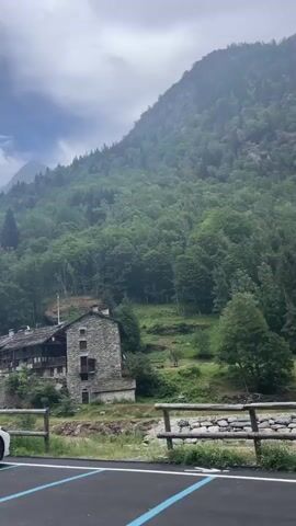 The video shows a panoramic view of a mountainous region with lush green forests. In the foreground, there is a parking lot with several cars. One car is a white sedan, another is a silver pickup truck with a rooftop tent, and a third is a grey minivan. A stone building with multiple windows and a dark roof is visible on the mountainside. In the background, the mountain slopes are covered with dense trees, and the sky is partly cloudy. The camera pans across the scene, showcasing the natural beauty of the area and the parked vehicles.