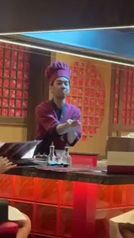 A medium shot shows a man in a maroon chef's uniform and hat reaching across a counter towards a man sitting on the opposite side. The man at the counter has his hands up near his chest as the chef reaches out. The counter is red, with a clear divider behind it that is framed in red and black. A second chef can be seen in the background, also dressed in maroon. The man seated at the counter is wearing a white shirt and patterned shorts. Other people can be seen at the counter on either side of the two main subjects. The restaurant has an asian decor.