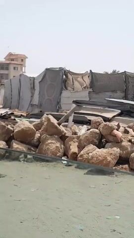 The video shows a series of tent-like structures in an outdoor, sandy environment. Initially, a pile of rocks and metal scrap obstructs the view. As the camera moves, a young girl with a backpack walks past. The video then reveals more tents, and finally, a silver car with a missing front bumper is partially visible, surrounded by more metal scrap. The scene suggests a makeshift or temporary settlement with possible debris or salvage materials nearby.