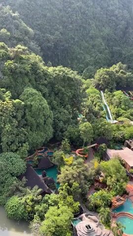 This aerial video shows a lush green jungle surrounding a resort with several water slides and swimming pools. The water slides are various colors, including white, orange, and red, and are set against the backdrop of dense foliage. Swimming pools with turquoise water are visible, some with lounge chairs and umbrellas. Buildings with dark roofs are scattered throughout the resort area. The scene appears to be an outdoor recreational facility nestled within a natural, mountainous landscape. The video pans slightly, offering a dynamic view of the entire area.