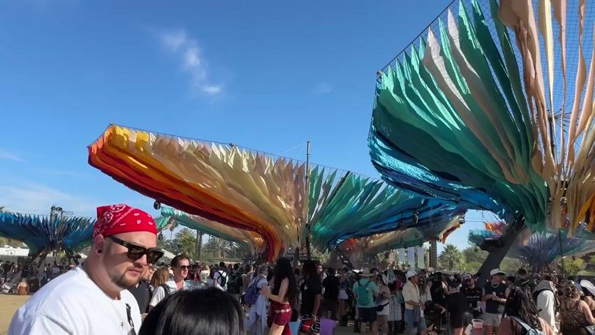 The video captures a lively outdoor festival scene under a bright, clear blue sky. Numerous people are gathered in a large, open, dusty area, walking, mingling, and sitting on the ground. Overhead, several massive, colorful art installations dominate the view, appearing as giant, fan-like structures made of flowing fabric panels in vibrant shades of red, orange, yellow, green, blue, and purple. Black speaker systems are visible, attached to these art pieces, indicating music is playing. Attendees are dressed in casual, light clothing, with many wearing sunglasses and hats (including baseball caps and a cowboy hat). The overall atmosphere is relaxed and festive. Distant mountains are visible in the background, confirming the setting in Indio, United States of America. The time of day is sunny, likely mid-day, with excellent visibility.