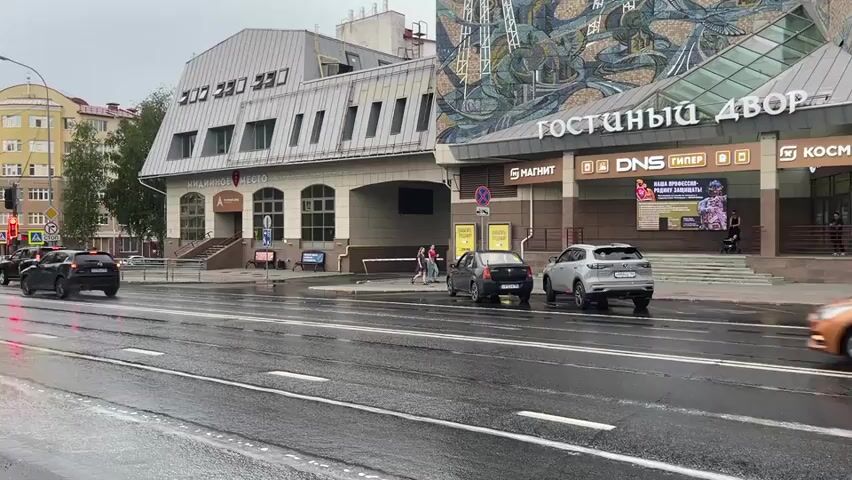 A street scene on an overcast day with a wet road. Cars are driving on the road, and some are parked on the side. In the background, there is a large building with signage that reads "ГОСТИНИЙ ДВОР" (Guest House) and other store names like "МАГНИТ" (Magnit) and "DNS ГИПЕР" (DNS Hypermarket). There are a few pedestrians walking on the sidewalk. The weather appears to be rainy, judging by the wet road and the overcast sky.