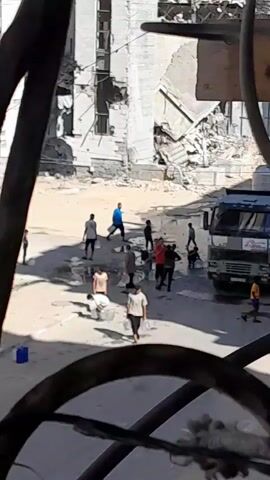 The video shows a group of people, including children and adults, in an outdoor area that appears to be damaged or under construction. Several individuals are carrying large translucent containers, possibly filled with water. The environment is dusty and the ground is uneven. Some people are walking, while others appear to be standing or crouching. There are also some blue containers visible on the ground. The overall impression is one of people engaged in collecting or transporting resources in a difficult setting.