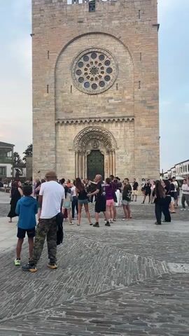 The video begins facing a large stone building with a rose window and an arched doorway. A crowd of people are gathered in a cobblestone plaza in front of the building. The camera pans slightly to the left, revealing more of the plaza and other buildings. A stage with musicians is visible in the background, with a sign that reads "DOMINGOS FOLCLORICOS DE PORTOMARIN." Several children are standing in the plaza, some watching the musicians, others looking towards the camera. The sky is overcast, suggesting it is either early morning or late afternoon. The overall atmosphere appears to be that of a community event or festival.