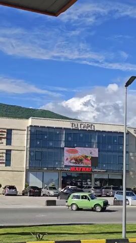 The video shows the exterior of a building with the name "ТцГАРТI" on top and the word "МАГНАТ" in large red letters on the front, which also says "Супермаркет" in smaller letters. The word "Афатәтырҭа" is above the word "МАГНАТ". The building is surrounded by a parking lot with several cars. The sky is blue with some white clouds, and there is a mountain in the background. There is an advertisement on a large video screen attached to the building. The camera zooms in on the store.