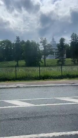 A video captures a view of a large, light-colored building with multiple levels and a domed roof, set on a grassy hill behind a dark fence. The building appears to be surrounded by trees. In the foreground, a wet asphalt road with white lane markings is visible. The sky is cloudy. The camera appears to be moving, as the perspective shifts slightly throughout the short clip.