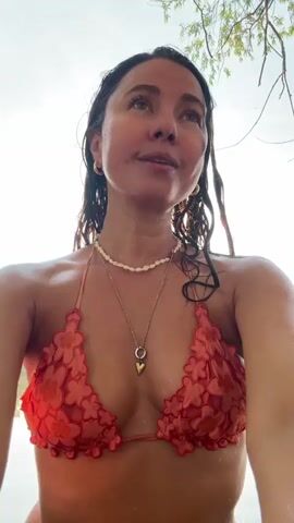 A woman with wet hair and a red floral bikini top looks into the camera from a body of water. She is wearing two necklaces, one with a white beaded strand and another with a gold pendant. In the first frame, trees are visible in the background. In subsequent frames, the water and the sky are visible, with the sun glinting off the water. The woman closes her eyes in the last frame.