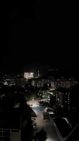The video shows a nighttime aerial view of a city. The sky is dark, with some distant lights visible on a hillside. In the foreground, there are several buildings, including apartment complexes and a taller, brightly lit building in the center. A street with cars parked along the side is visible in the middle ground. Trees are scattered throughout the scene, and streetlights illuminate parts of the area. The overall impression is of a quiet, illuminated city at night.