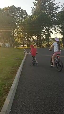 Two children are riding bicycles on a paved path. The child in front is wearing a red shirt and hat, and the child behind is wearing a white shirt and pink hat. The path is lined with grass and trees. The sun is setting, casting a warm glow on the scene. The child in front lifts one leg off the pedal and extends it outwards while riding.