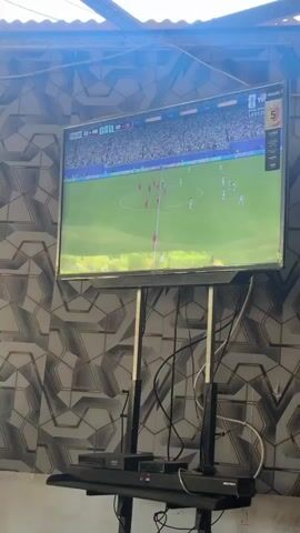The video shows a television screen displaying a soccer match. The game is being played on a green field with spectators in the background. The scoreboard on the screen shows the time as 11:32, and the score is 2-1. One team is wearing red uniforms, and the other is wearing white uniforms. The television is mounted on a stand, and there are some cables visible below it. The wall behind the television has a patterned design.
