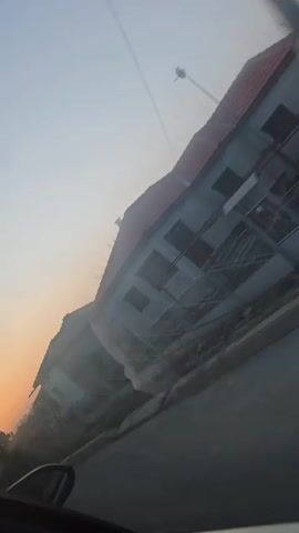 The camera is inside a car moving along a paved road. On the right side of the road are houses and trees. The houses are mostly gray with orange tile roofs. The trees are bare, except for some evergreens. The sun is setting in the distance, creating a warm orange glow in the sky. The video is somewhat shaky due to the movement of the car.