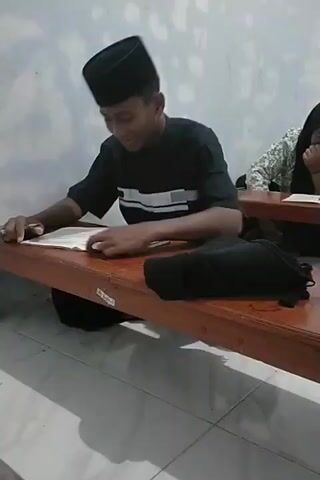 The video shows two young people sitting at wooden desks, reading. The person in the foreground, a young male wearing a black peci (a type of cap), is looking down at a book open on the desk in front of him. He is wearing a black t-shirt with white stripes across the chest. He appears to be speaking or reacting to the text. To the right, a young female wearing a hijab is also visible, looking down at her book and smiling. The environment appears to be a classroom or study area, with simple walls and tiled flooring. A black bag is visible on the bench between the two individuals.