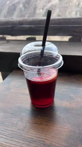 The video begins with a close-up of a red beverage in a plastic cup with a lid and black straw, sitting on a wooden table. The camera then pans upward to reveal a mountainous landscape in the background, seen through a window or opening. The mountains are mostly bare, with some trees. The sky is overcast, with patches of blue. A wooden railing is visible in the midground. The camera continues to pan to the right, showing more of the mountain range and a rural scene with a green tarp structure and a few scattered trees.