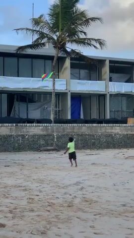 A young boy in a neon green shirt and dark shorts stands on a sandy beach and flies a kite. The kite, which is multicolored, flies in the sky above and to the right of the boy. Behind the boy and the beach is a multi-story building with large glass windows and a tall palm tree. The building appears to be under construction, with some windows covered and scaffolding visible. The sky is partly cloudy and the lighting suggests it is either early morning or late afternoon. The scene is calm and peaceful.