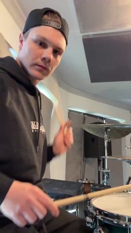 A young man wearing a black hoodie and a backwards black baseball cap is playing the drums in a recording studio. The drum set is a dark green color, and the man is using wooden drumsticks. The studio has soundproofing panels on the walls and ceiling, and a doorway is visible in the background. The man is looking at the camera as he plays, and appears focused.