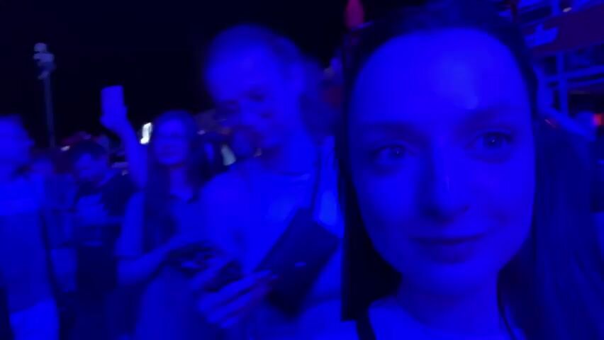 The video shows a group of people at an outdoor event, likely a festival or concert, under a nighttime sky. The scene is illuminated by vibrant blue lighting, casting a cool hue over everything. In the foreground, two young women are prominently featured, taking a selfie. The woman on the right, closer to the camera, smiles broadly, her teeth visible. The woman on the left, wearing glasses, also smiles and looks towards the camera. They are surrounded by other attendees, some blurred in the background, suggesting a lively crowd.

One person in the foreground is wearing a t-shirt with the text "IRON MAIDEN" visible. Behind the main subjects, a banner or sign with some indistinct text in red and yellow colors is partially visible, along with what appears to be a stage or elevated platform. The overall atmosphere seems energetic and festive, with people gathered closely together. The presence of a t-shirt referencing a popular band and the crowded setting suggest a music event. The time of day is clearly night, indicated by the darkness of the sky and the artificial lighting. The location is identified as Kragujevac, Serbia, though this is not visually evident from the provided frames alone. The emotions conveyed are primarily joy and excitement, as expressed by the smiles of the two women and the general dynamic of a crowd at an event.