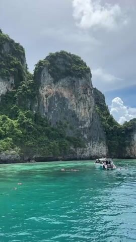 A drone shot pans across a body of water, revealing towering cliffs covered in lush green foliage. In the foreground, the turquoise water is clear and calm. A white speed boat with several people on board is anchored near the shore, while others swim and float in the water. In the distance, other boats and islands can be seen. The sky is a mix of blue and white clouds.