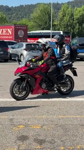 Two individuals, clad in full motorcycle gear including helmets, are seated on a vibrant red sportbike. The rider in front wears a white helmet and a black and red jacket, while the passenger behind them has on a black helmet and a blue and white jacket, with a black backpack strapped on. The motorcycle is parked on an asphalt surface marked with yellow lines, indicative of a parking lot or rest stop.

In the background, several vehicles are visible, including cars and large trucks. One truck prominently displays the text "LAURITZEN LOOKING FOR GREENER WAYS." Another truck has the word "SAMEG" on it. To the right, a van with "Dental" visible on its side suggests a commercial presence. Further back, a sign advertising "Joy of Outlet Shopping" and "Franciacorta Designer Village" is mounted on a blue stand. Text on the sign also includes "OLTRE 190 BRAND CON RIDUZIONI DAL 30% AL 70% TUTTO L'ANNO" and "OVER 190 BRANDS WITH 30-70% REDUCTIONS ALL YEAR ROUND." It also states "FranciacortaDesignerVillage.com" and "AUTORSTRADA / HIGHWAY A4 > USCITA/EXIT OSPITALETTO". The setting appears to be a sunny day, possibly midday given the bright light and shadows, with a hilly, tree-covered landscape in the distance. The scene suggests a stop during a motorcycle journey, perhaps at an outlet mall or service area.