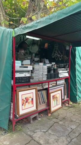 The video shows a flea market stall under a green and red awning. The stall is filled with vintage items including stacks of CDs, vinyl records, vintage audio equipment, and framed artwork. The items are neatly arranged on shelves within the stall. The stall is situated outdoors with trees visible in the background. The camera zooms in on the stall's contents.