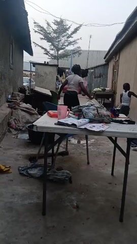 The video shows a woman and two children in what appears to be a narrow outdoor space between buildings. The space has a concrete floor and is cluttered with various items, including clothes, buckets, and miscellaneous debris. A white folding table is in the foreground, covered with a notebook, clothing, and other objects. The woman walks up to the table carrying a white object and begins to clean it. One child stands near the table while the other child stands near a doorway to the right. Clothing is hung on a clothesline in the background between two buildings. A tree is visible behind the buildings.  The overall scene suggests a modest living environment.