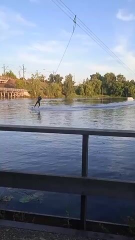 A person is wakeboarding on a body of water. They are being pulled by a cable. There is a bridge in the background. The water is calm and there are trees on the shore. The sky is blue with a few clouds.