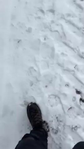 The video shows a person walking in the snow. The camera angle is low, showing the person's black boot and pants. The snow is white and has footprints in it. The camera then pans up to show a snow-covered path leading up a hill. There is a stone wall on the left side of the path and bare trees on either side. The sky is cloudy and grey.