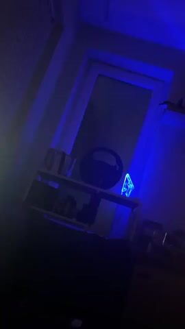 The video shows a dimly lit room with blue ambient lighting. On a shelf, a black steering wheel is visible, along with a small, translucent blue object. A television screen displays an image of silhouetted figures walking. A black gaming chair is positioned in front of the TV. There is a plant in a pot and a tennis racket hanging on the wall.