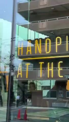 The video shows a building in an urban setting. Initially, a bright yellow neon sign reading "HANDPICKED ROASTER" is visible through a glass window. The camera pans up, revealing a building with multiple stories. The camera angle shifts to show street signs bearing a star and the letter "R", along with the words "TEAVANA" and "ARRIVIAMO". The signs are mounted on a pole, and a building is visible in the background.