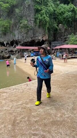 A person wearing a blue shirt with black and purple accents, black pants, a wide-brimmed hat, and yellow shoes is standing on a sandy beach. They are holding a black object in their right hand and a red object in their left hand. The person is looking upwards and to their right. In the background, there is a body of water, a sandy beach, and a large rock formation with vegetation. There are also several structures with red roofs and people walking around them. The weather appears to be overcast.