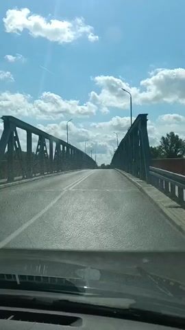 The video is filmed from inside a vehicle, looking out through the windshield as it drives across a bridge. The bridge is made of dark metal with a repeating pattern of vertical supports and diagonal beams. The road surface is asphalt with white lines marking the lanes. On either side of the road are metal railings, and beyond the railings, trees and a body of water are visible. The sky is blue with scattered white clouds, and sunlight is reflecting off the wet road. Streetlights are visible at intervals along the bridge. The vehicle appears to be moving at a moderate speed.