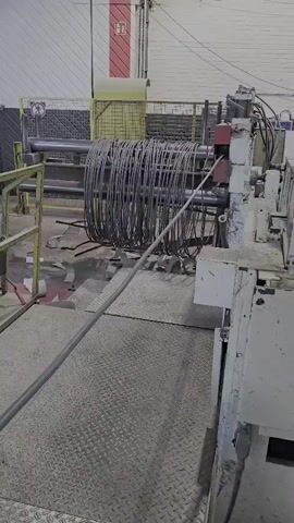 A metal wire is being fed into a machine in a factory. The wire is being coiled onto a large cylindrical drum. The machine appears to be automated, with no people visible in the frame. The environment is a typical industrial setting, with concrete floors and metal structures.