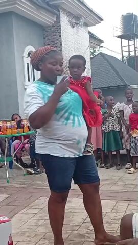 The video captures an outdoor gathering of children and adults in Onicha, Nigeria. The central figure is a woman wearing a tie-dye t-shirt, dark shorts, and a patterned head covering. She is actively engaged with the children, speaking or singing while holding a small, clear object, possibly a bottle or a makeshift microphone. At the beginning, she carries a baby, which she later passes to another woman wearing a blue and white dress.

A large group of children, ranging from toddlers to older elementary school age, are gathered around, attentively watching the woman. They are dressed in various casual outfits, with one child briefly visible wearing a white t-shirt with "GUCCI" printed on it.

In the background, there is a table laden with numerous colorful drinks in plastic bottles or pouches, suggesting a refreshment station. A light grey and stone-clad house with arched windows is visible behind the children. Further back, a tall water tank on a stand can be seen against an overcast sky. The ground is paved, and there are trees and foliage around the perimeter, indicating a residential or community setting. The atmosphere appears lively and engaging, with the children focused on the activity led by the woman.