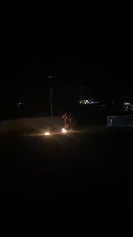 A person is performing a fire show at night. They are holding a device that is producing a large amount of sparks, which are flying outwards in a circular motion around the performer. There are also several fires burning on the ground around the performer. The background is dark, with some distant lights visible. The person is silhouetted against the sparks and flames. The ground appears to be dirt or sand.