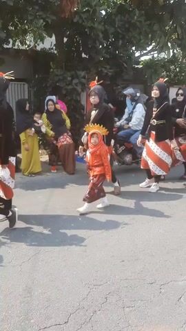 A group of people are walking down a street outdoors in what appears to be a parade or procession. The people in the foreground are mostly children wearing traditional clothing, including ornate headwear and brightly colored outfits. Some adults in black clothing and headscarves are walking alongside them, appearing to supervise or participate in the event. There are trees and buildings visible in the background, and the ground is paved. The lighting suggests it is daytime.