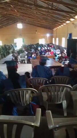 A wide shot shows a gathering in a hall with a wooden roof. In the foreground, several people wearing academic regalia, including blue gowns and caps, are seated. The majority of the attendees are seated in rows facing a stage area at the front of the hall. Some people are standing. The hall appears to be filled with a large crowd. A woman in a patterned dress is standing and appears to be speaking or being presented. The environment is brightly lit, possibly by natural light from windows or overhead lighting.