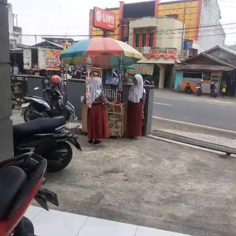 The video shows an outdoor scene, possibly a street or sidewalk, with several motorcycles parked on the left side. In the center, two young individuals wearing school uniforms, consisting of a maroon skirt and white hijab, are standing next to a small, colorful umbrella-covered cart filled with items for sale. One individual appears to be interacting with the cart, possibly as a vendor or customer, while the other stands nearby. Behind the cart, there are buildings, including a shop with a red sign that reads "ELIJANTA." The overall environment appears to be a commercial area with moderate foot traffic.