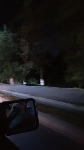 A first-person view from inside a vehicle driving at night. The side mirror shows a hand forming a heart shape. The road ahead is illuminated by streetlights and the headlights of other vehicles. Trees and buildings are visible along the roadside. A speed limit sign indicating 60 is visible.
