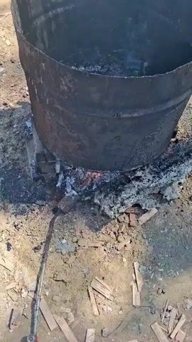 A large metal barrel is positioned over a fire. The barrel is black and appears to be scorched. It contains a dark, bubbling liquid. The ground around the barrel is dirt and debris, with some scattered wood fragments. A long metal pole with a spatula-like end is being used to stir the contents of the barrel. The fire beneath the barrel is visible, with flames and glowing embers. The scene suggests a process of heating or melting a substance, likely asphalt or tar, for construction or road repair.