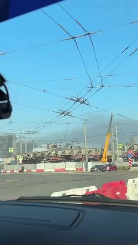 The video shows a view from inside a vehicle, looking out at a street scene. The sky is clear and blue. Overhead, a complex network of wires, likely for an electric transport system like a trolleybus or tram, crisscrosses the frame. In the foreground, the dashboard of the vehicle is visible. The road ahead is lined with temporary red and white barriers, indicating construction or roadwork. Several vehicles, including cars and what appears to be a tram or trolleybus, are present on the road. Construction equipment, including a yellow crane, is visible in the background, along with partially constructed buildings or bridges. Traffic signals and road signs are also visible, including a red and white chevron sign indicating a turn. The overall impression is of an urban environment undergoing significant development or repair.