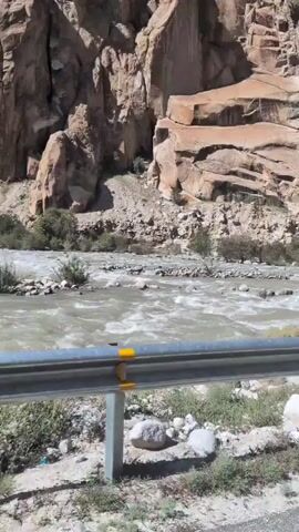 The video shows a view from a moving vehicle. In the foreground, a silver metal guardrail with yellow and black reflective stripes is visible. Beyond the guardrail, there is a rough terrain with stones and some sparse vegetation. A river with murky, fast-flowing water flows through the middle ground. Large, light brown rock formations with visible striations and geological layers dominate the background. The sky above is a clear, bright blue. The overall scene depicts a mountainous river valley.