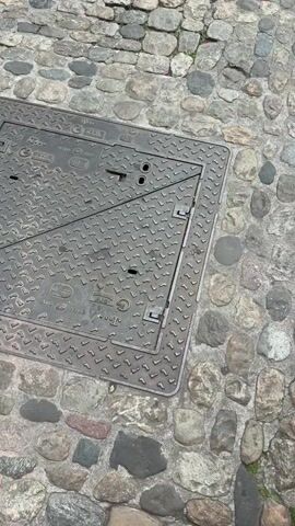 This video captures a walk through Freiburg im Breisgau, Germany. The scene opens with a close-up of a cobblestone street and a metal manhole cover, suggesting an urban setting. Two pairs of feet, one in white sneakers and another in black flip-flops, are visible at the bottom of the frame, indicating someone is filming from a low, personal perspective.

The perspective then shifts to show the entrance of a large, ornate church, likely Freiburg Minster, constructed from reddish-brown stone. The architecture features Gothic elements, including pointed arches and intricate carvings. Several people are walking around the plaza in front of the church. The weather appears to be partly cloudy, with visible blue sky and white clouds.

The camera then pans up to reveal the full height of the church's tower, a striking spire reaching towards the sky. A large, circular clock with a red and blue face is a prominent feature on the tower. Scaffolding is visible on one side of the tower, suggesting ongoing maintenance or restoration work. The overall impression is one of historical grandeur and a lively public space. No legible text is visible in the video.
