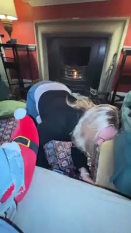 A woman on her hands and knees retrieves two small bottles of alcohol from under a sofa. She is wearing jeans and a black sweater. Someone is sitting on the sofa with their feet up, wearing jeans and novelty Santa-themed slippers. The room has a fireplace and a rug with an intricate pattern. The walls are red.