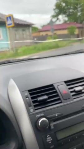 The video shows a view from inside a car, looking out through the windshield on a rainy day. The windshield wipers activate to clear the rain. The camera captures the dashboard of the car, showing the radio, hazard light button, and a glimpse of the steering wheel. Outside the car, there's a narrow paved road running through a residential area.  Houses are visible on either side of the road, characterized by traditional architecture and surrounded by trees. Power lines are also strung overhead.  In the distance, a yellow bus drives down the road. The sky is overcast, contributing to the gray, muted color palette of the scene.