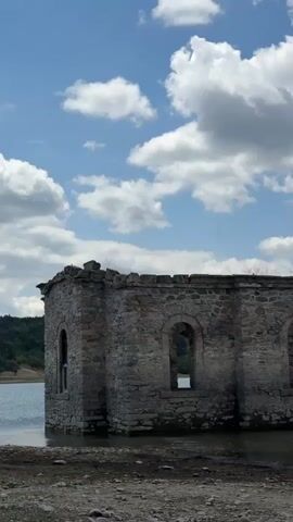 A stone structure, partially submerged in water, stands under a cloudy sky. The structure appears to be a ruin of a building, possibly a church, with arched windows and a cross visible on its roof. The water level is low, exposing a rocky shoreline. In the distance, hills and a body of water are visible. The scene is static, with no people or animals present.