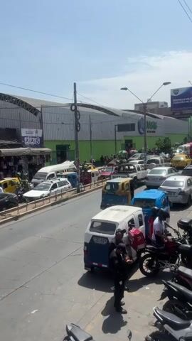 The video shows a busy street in a city, likely during the day, with a significant amount of traffic. Cars, motorcycles, and auto rickshaws are tightly packed together. There are a few individuals, including people on motorcycles wearing delivery backpacks, and what appear to be police officers standing near a vehicle. Buildings line the street, one with the word "OASIS" visible on its exterior. Above the street is a large billboard advertising a health product. The sky is bright and sunny, adding to the overall impression of a bustling urban environment.