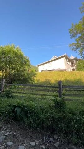 The video captures a landscape view with a house on a hill. The house is light-colored with an ornate balcony. A wooden fence runs across the foreground. The area is surrounded by greenery, including trees and grass, with a dirt road leading away from the scene towards mountains in the distance. The sky is clear and blue.